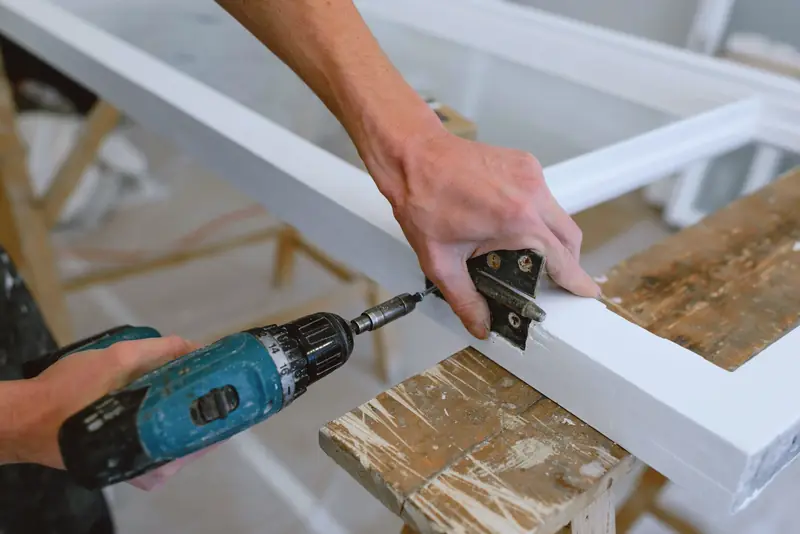Hands fitting a hinge to a white door frame with a cordless drill